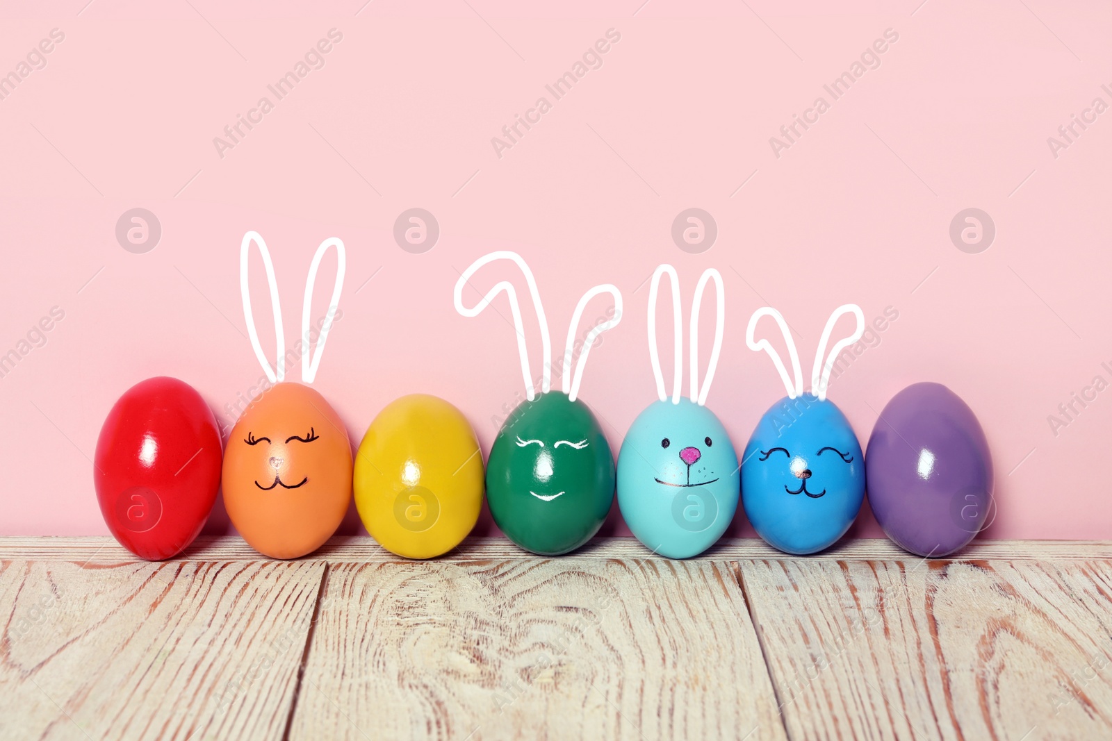 Image of Several eggs with drawn faces and ears as Easter bunnies among others on white wooden table against pink background