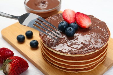 Photo of Eating tasty pancakes with chocolate paste and berries with fork on white wooden table, closeup