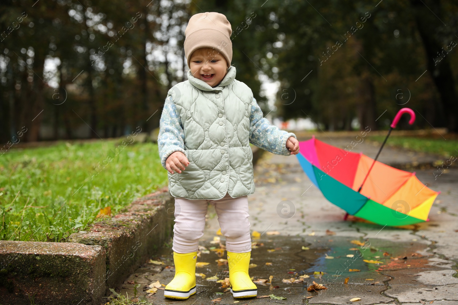 Photo of Cute little girl having fun in puddle near colorful umbrella outdoors