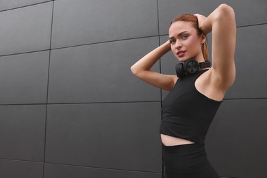 Photo of Beautiful woman in stylish gym clothes with headphones posing near dark grey wall on street, space for text