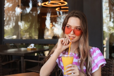 Happy young woman with heart shaped glasses and juice at cafe. Space for text