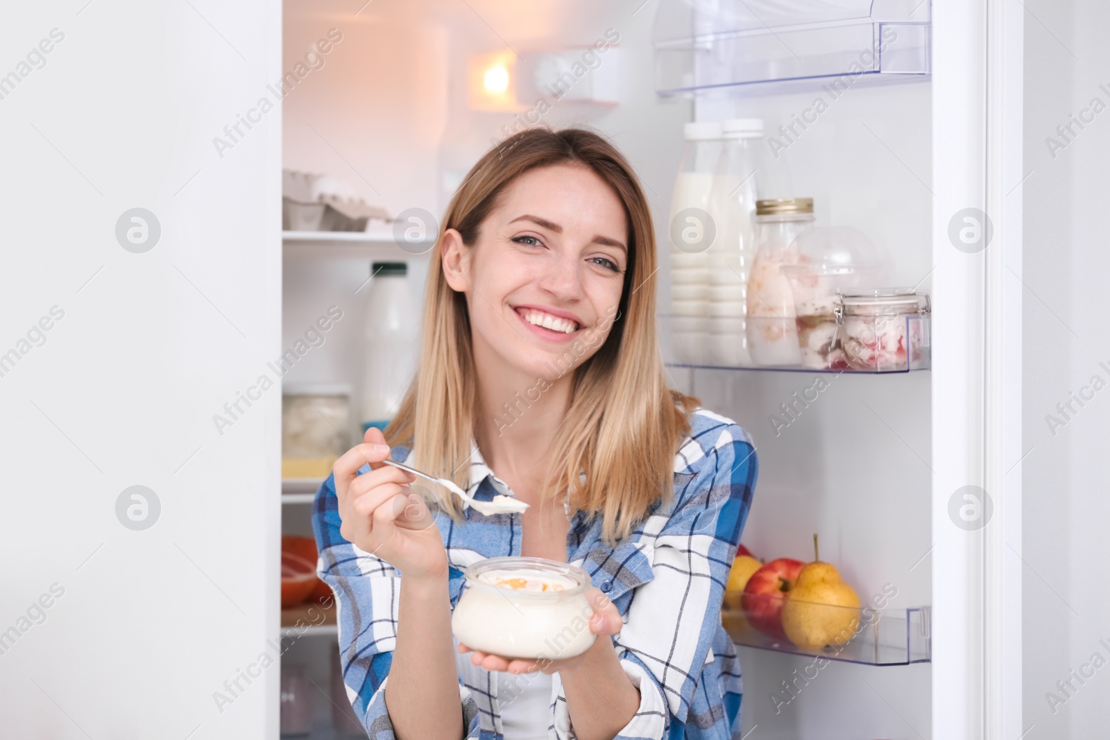 Photo of Young attractive woman with tasty yogurt near fridge