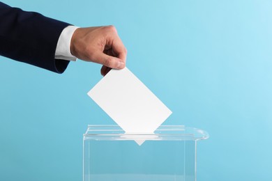 Man putting his vote into ballot box on light blue background, closeup