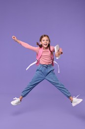Photo of Happy schoolgirl with backpack jumping on violet background