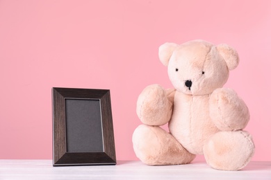Photo of Photo frame with space for text and adorable teddy bear on table against color background. Child room elements