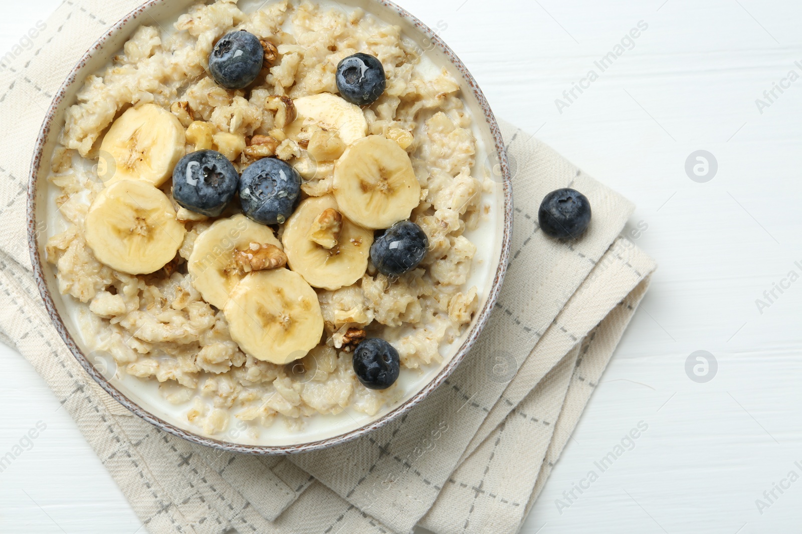 Photo of Tasty oatmeal with banana, blueberries, walnuts and honey served in bowl on white wooden table, top view. Space for text