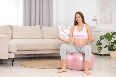 Photo of Pregnant woman meditating on fitness ball in room, space for text. Home yoga