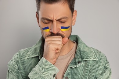 Sad man with drawings of Ukrainian flag on face against light grey background, closeup