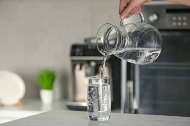Photo of Woman pouring water from jug into glass at wooden table in kitchen, closeup