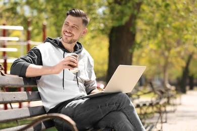 Portrait of young man with laptop and cup of coffee on bench in park