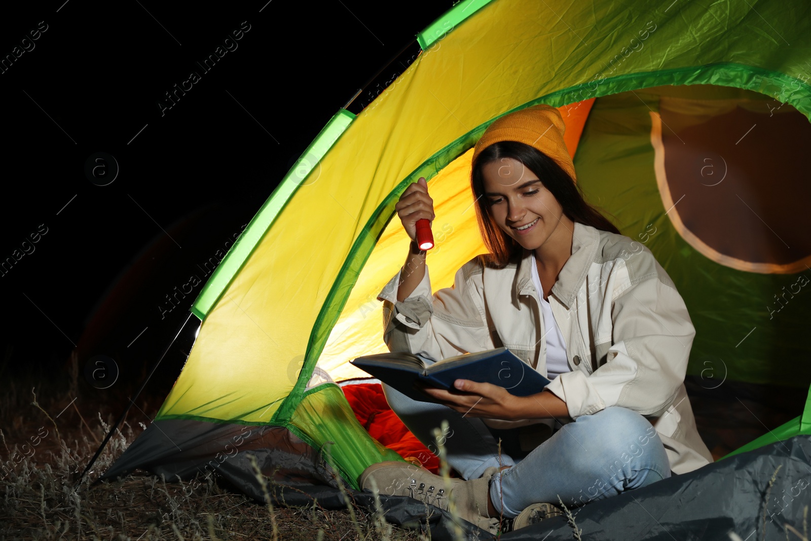 Photo of Young woman with flashlight reading book in tent at night