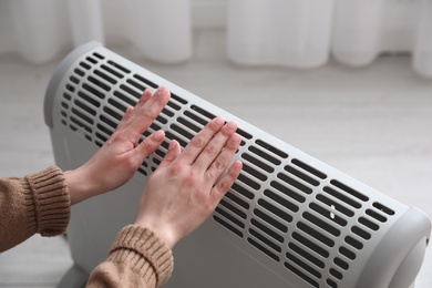 Woman warming hands near electric heater at home, closeup