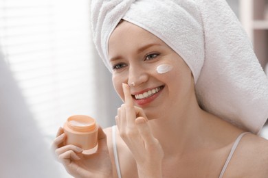 Photo of Smiling woman with freckles applying cream onto her face in bathroom