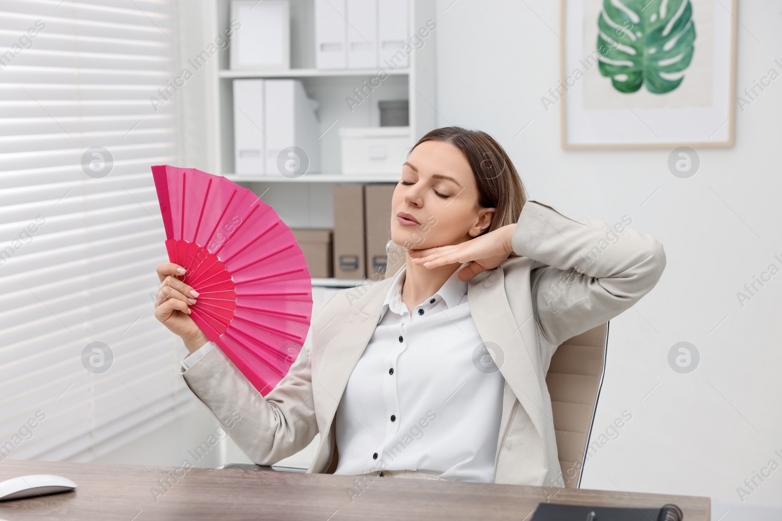 Photo of Businesswoman waving pink hand fan to cool herself at table in office