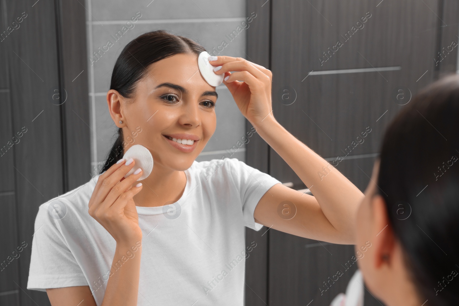 Photo of Young woman using cotton pads with micellar water near mirror in bathroom