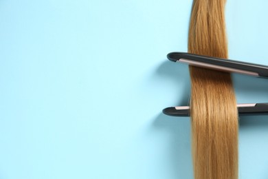 Modern straightener with blonde hair lock on light blue background, top view. Space for text