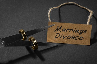 Photo of Scissors with wedding rings on blades and paper card Marriage Divorce against black background, closeup