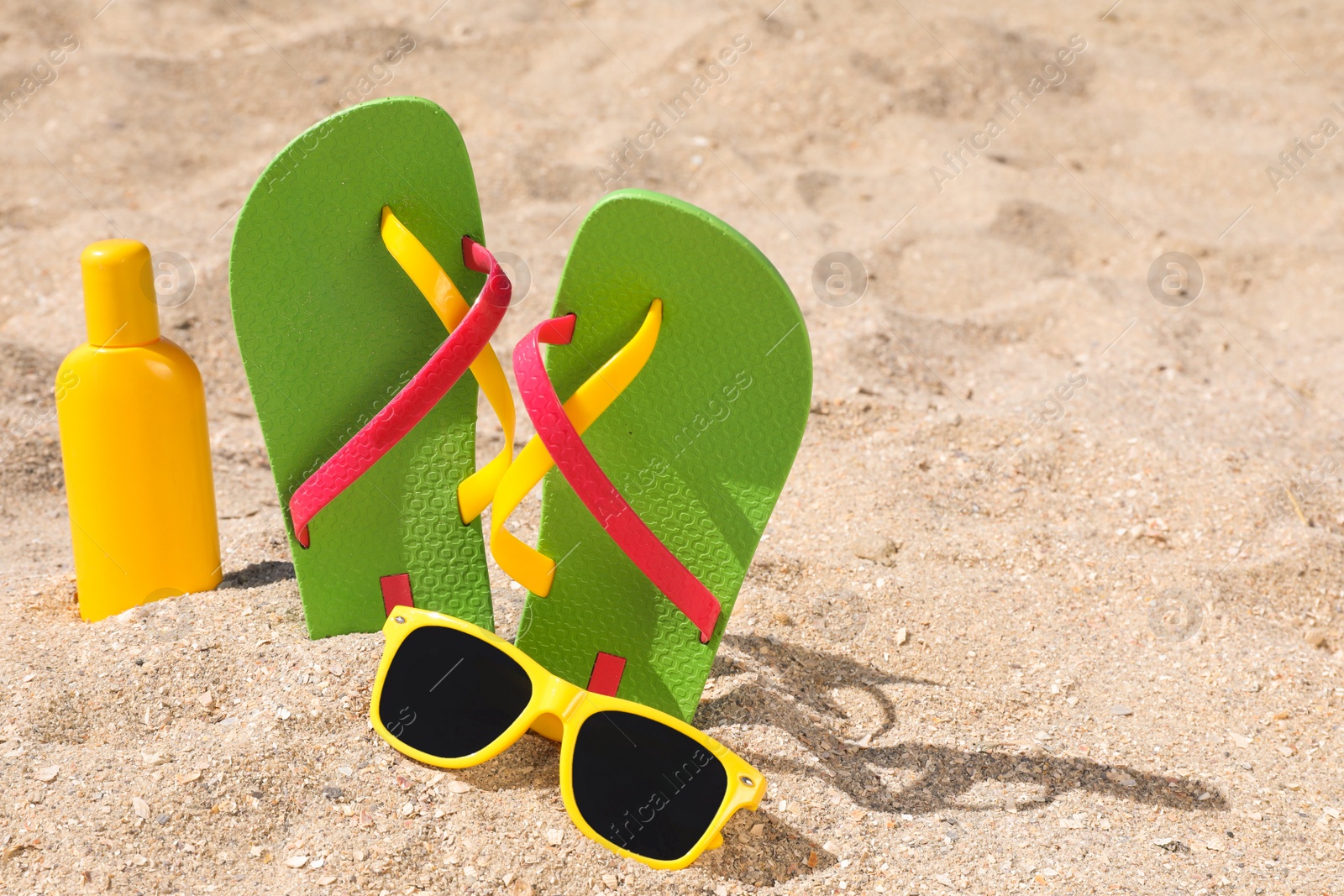 Photo of Stylish flip flops, sunglasses and sun protection cream on sand, space for text