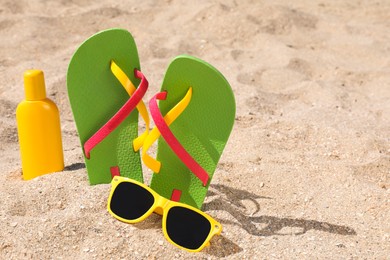 Photo of Stylish flip flops, sunglasses and sun protection cream on sand, space for text