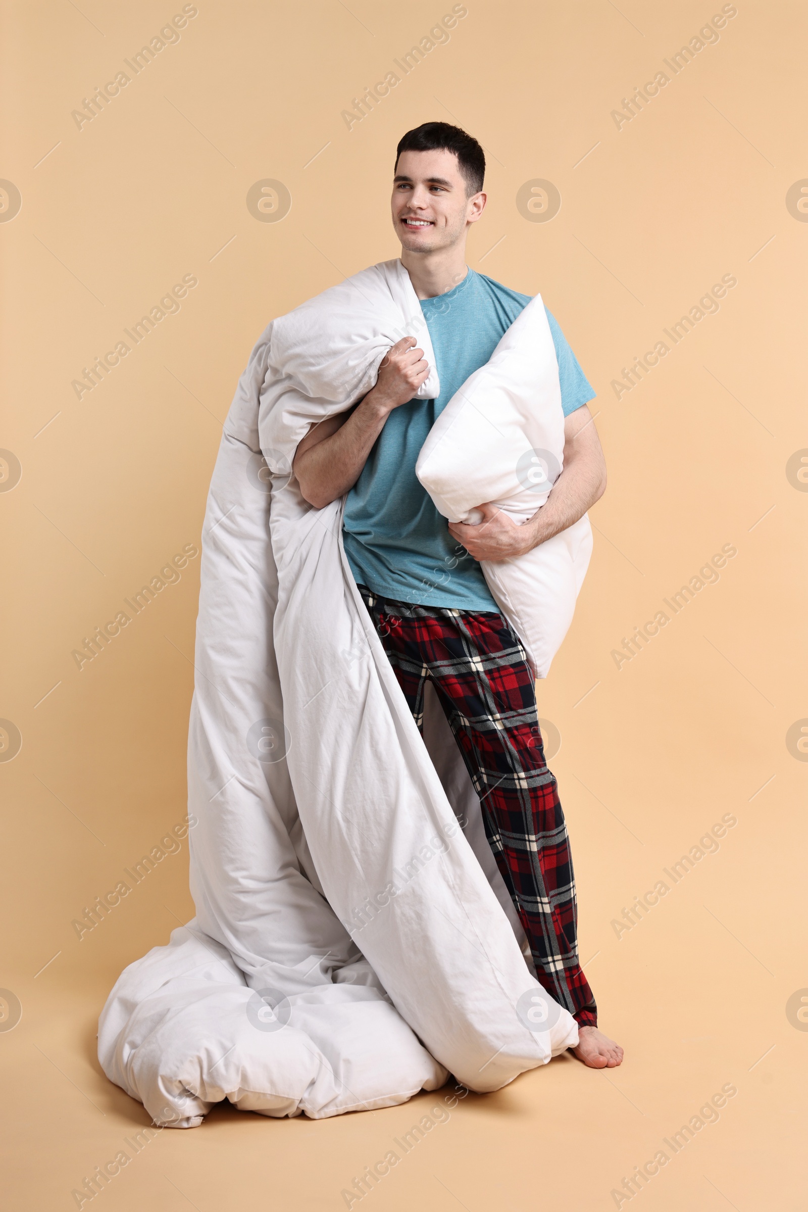 Photo of Happy man in pyjama holding pillow and blanket on beige background