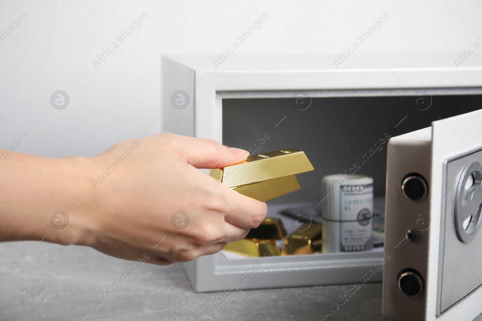 Photo of Woman putting gold bars into steel safe at light grey table, closeup