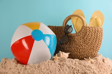 Photo of Colorful inflatable ball and straw bag with beach accessories on sand against light blue background