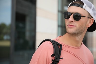 Handsome young man with stylish sunglasses and backpack near building outdoors, space for text