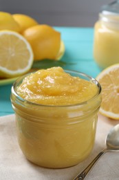 Photo of Delicious lemon curd in glass jar, fresh citrus fruits and spoon on table, closeup