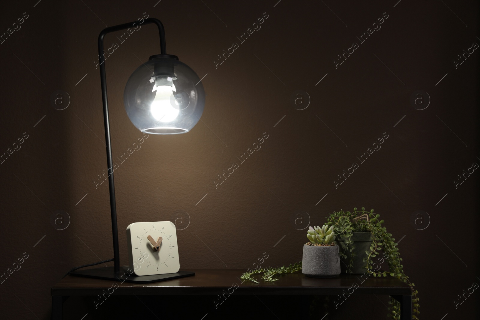 Photo of Stylish lamp, green plants and clock on table near brown wall indoors. Interior design