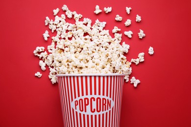 Photo of Overturned paper bucket with delicious popcorn on red background, flat lay