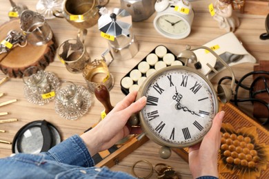Photo of Woman holding vintage alarm clock near table with different stuff, above view. Garage sale