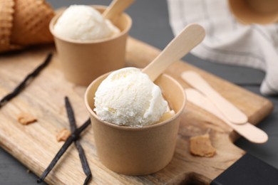 Photo of Paper cups with delicious ice cream and vanilla pods on wooden board, closeup