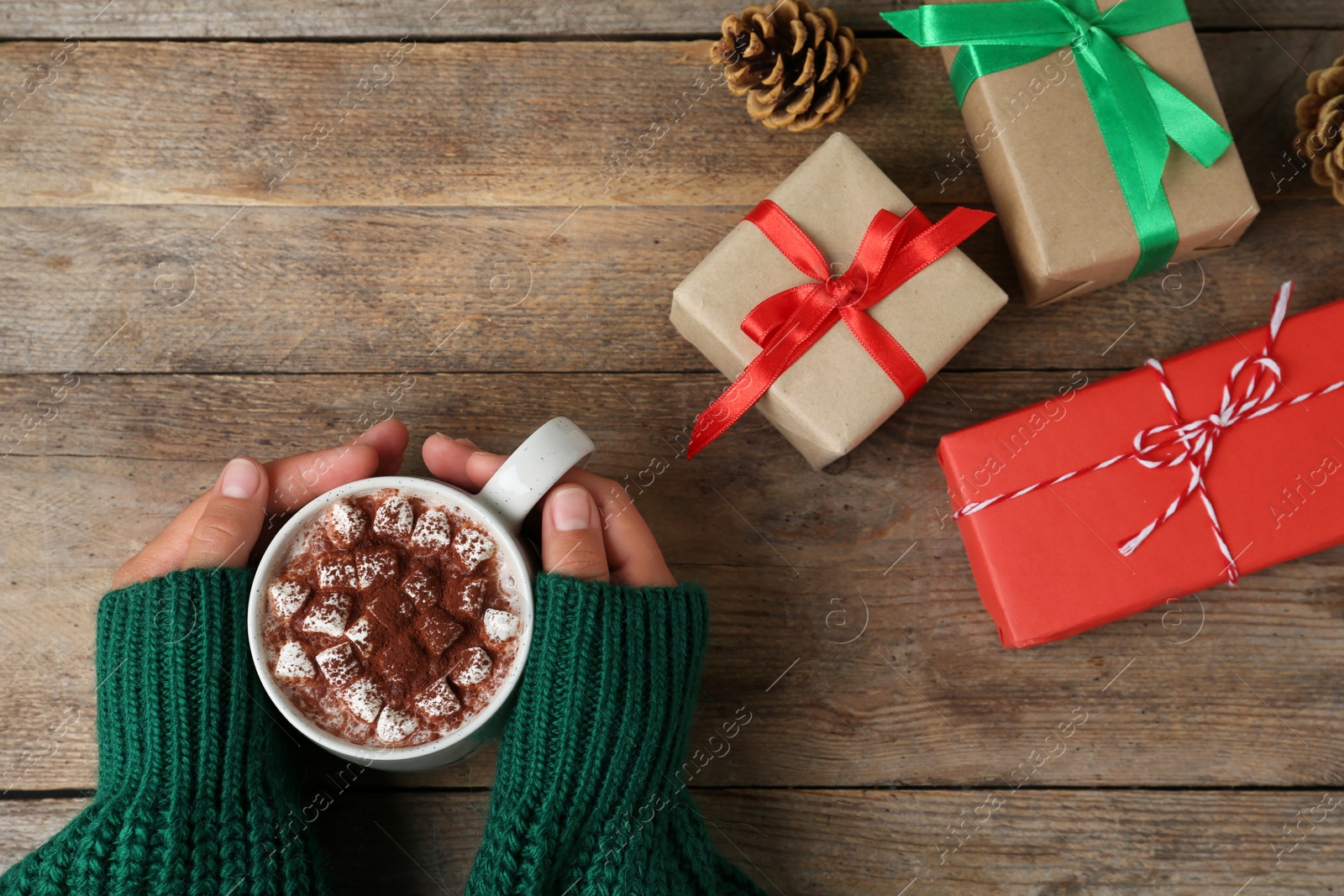 Photo of Woman holding cup of delicious cocoa drink at wooden table with Christmas presents, top view