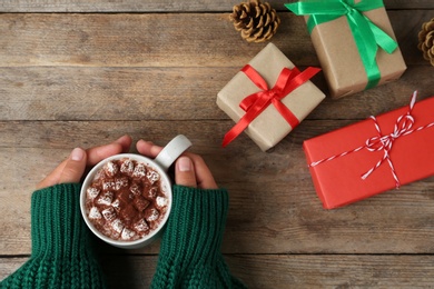 Woman holding cup of delicious cocoa drink at wooden table with Christmas presents, top view