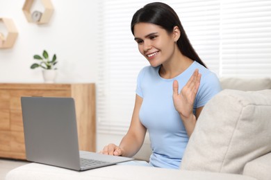 Happy young woman having video chat via laptop and waving hello on sofa in living room