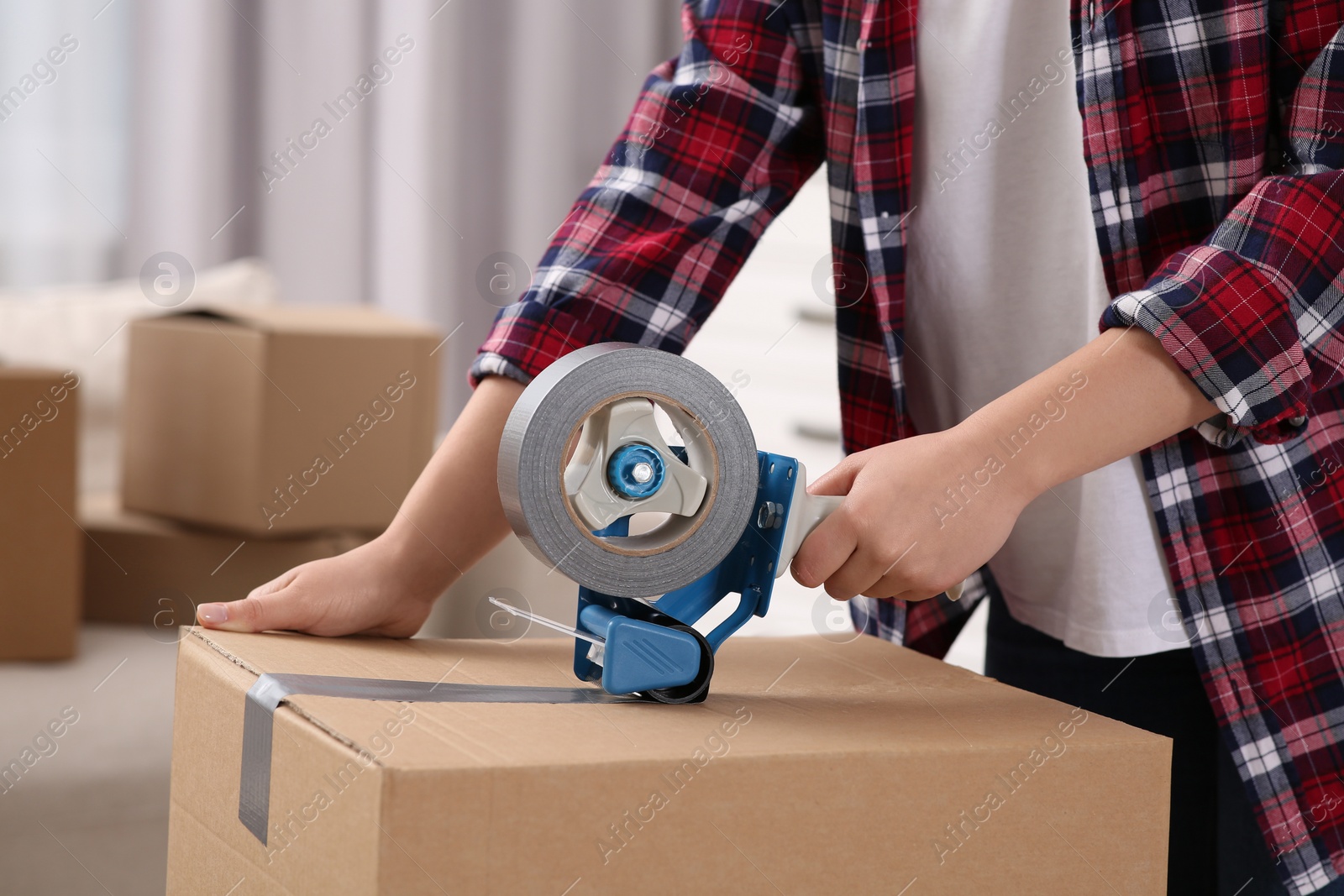 Photo of Woman applying adhesive tape on box with dispenser indoors, closeup