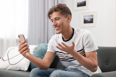 Photo of Happy young man having video chat via smartphone on sofa indoors