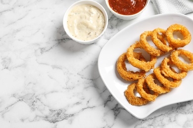 Photo of Homemade crunchy fried onion rings with sauces on marble table, top view. Space for text