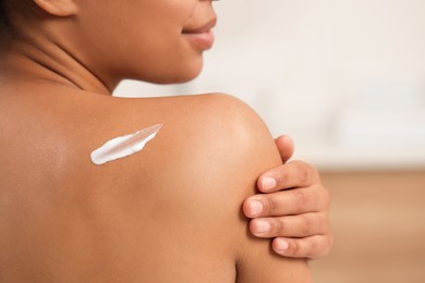 Young woman applying body cream onto back on blurred background, closeup