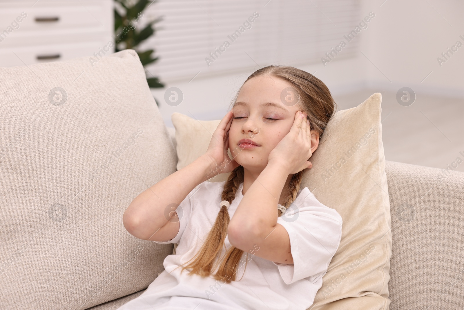 Photo of Little girl suffering from headache on sofa indoors