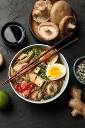 Photo of Bowl of delicious ramen, ingredients and chopsticks on black table, flat lay. Noodle soup