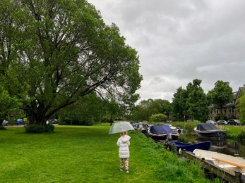 Photo of Little girl with umbrella walking near canal on rainy day, back view