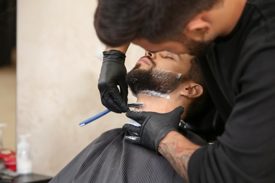 Professional hairdresser shaving client with straight razor in barbershop