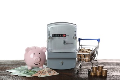Photo of Electricity meter, piggy bank and small shopping cart with money on wooden table against white background