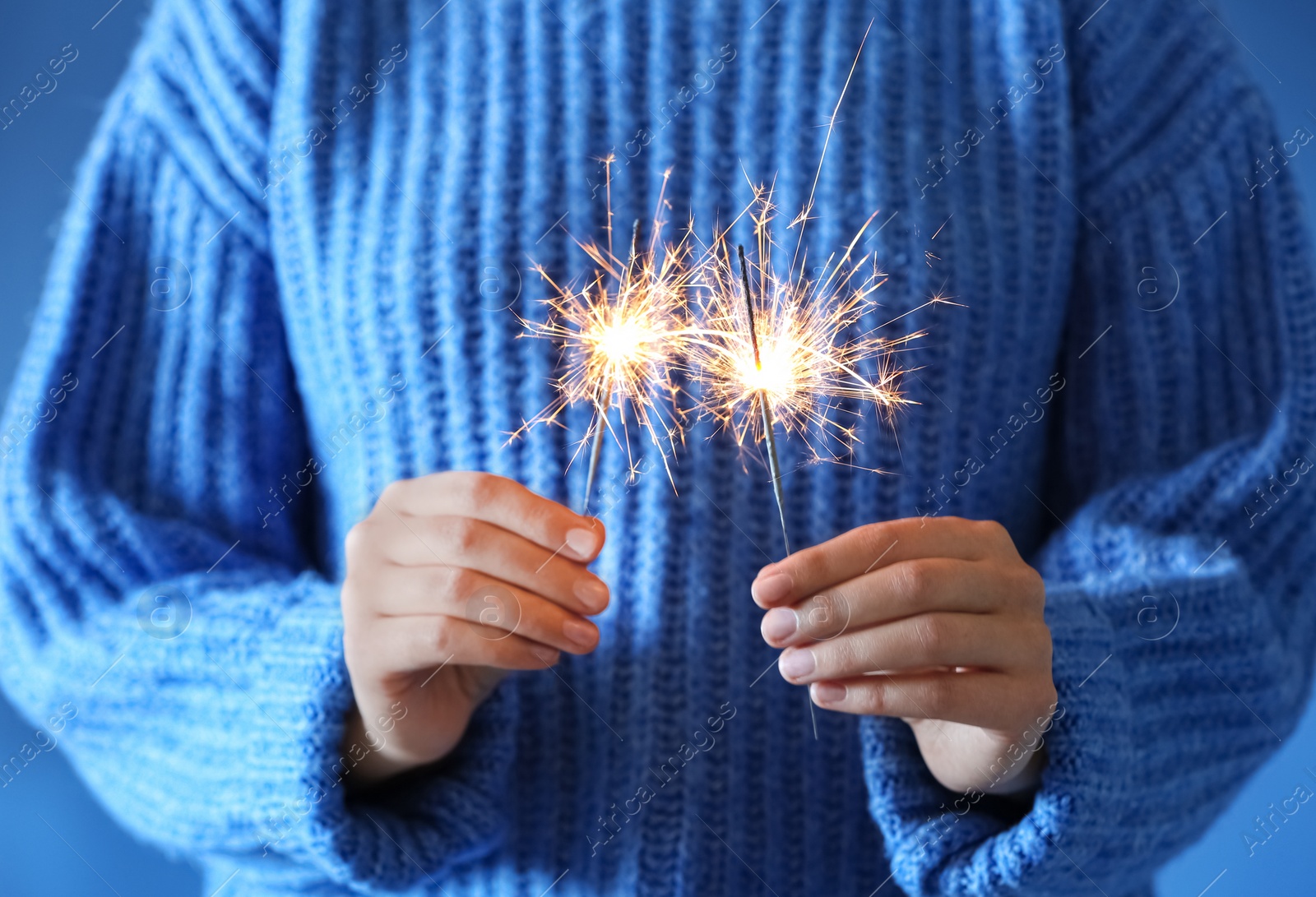 Photo of Woman holding burning sparklers on blue background, closeup