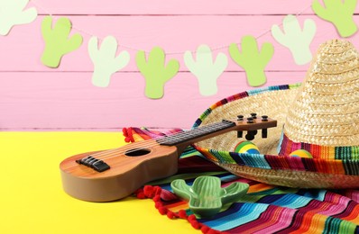 Photo of Mexican sombrero hat, poncho and ukulele on yellow table, closeup. Space for text