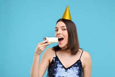Happy young woman in party hat eating cheesecake on light blue background