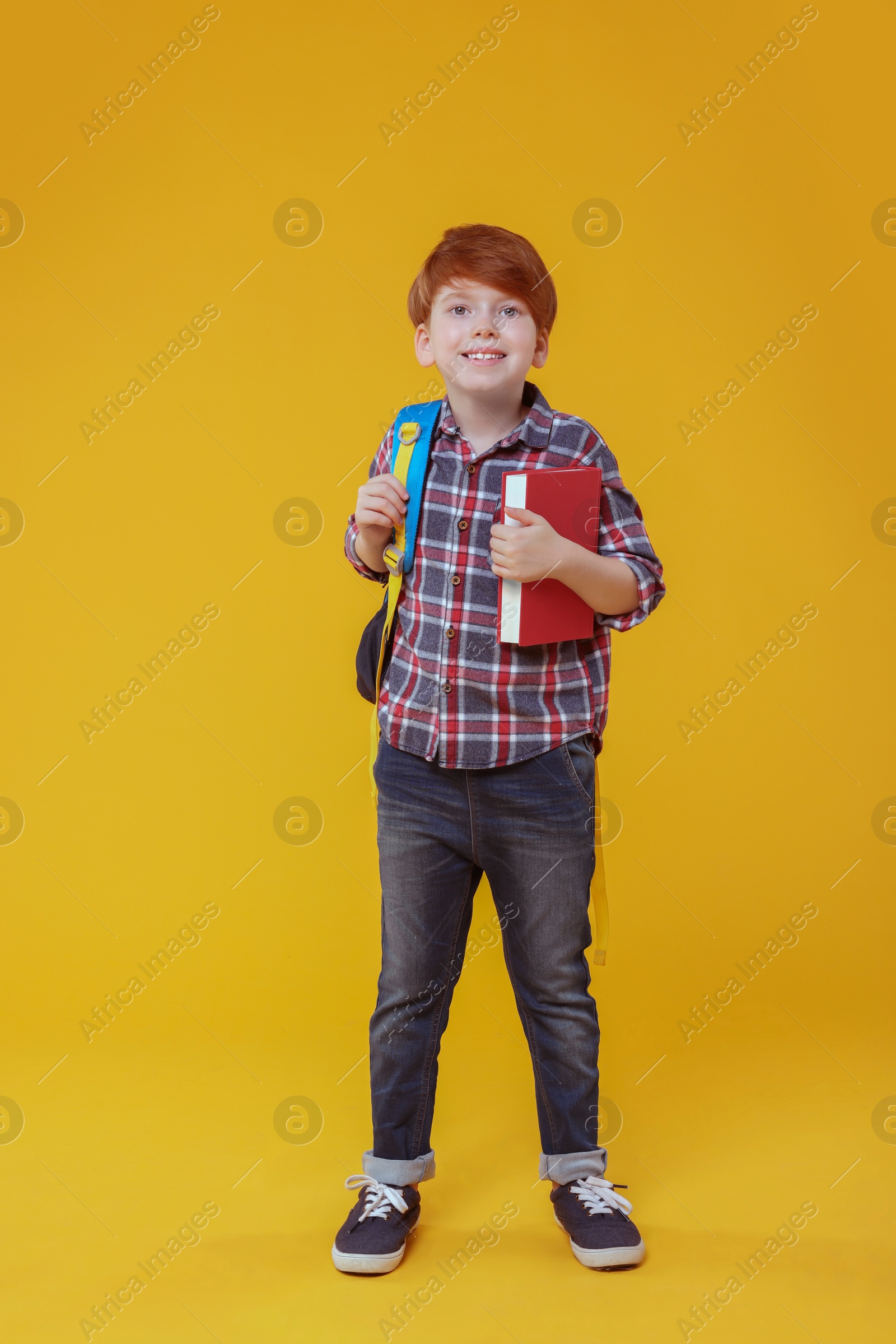 Photo of Smiling schoolboy with backpack and book on orange background