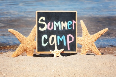 Photo of Little blackboard with text SUMMER CAMP and sea stars on sand near sea