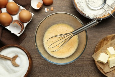 Photo of Beaten eggs, whisk and ingredients on wooden table, flat lay